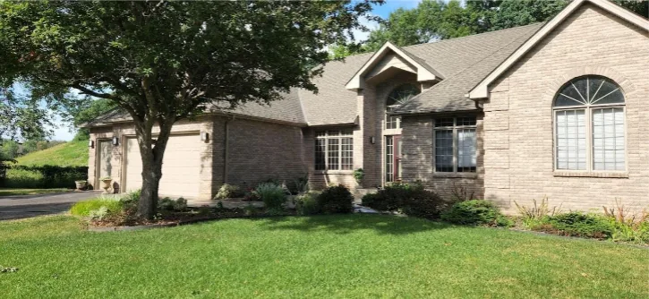 Front view of a single-story brick ranch-style home featuring tan brick veneer, arched windows, dark gray asphalt shingles, and mature landscaping under bright daylight.