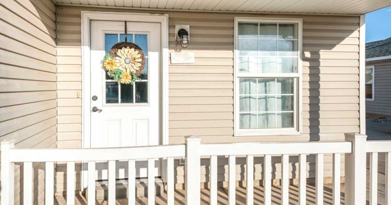Porch of a house with beige vinyl wood siding and railing. There is a white front door with wreath on top of the glass panels near the single hung window with curtain.