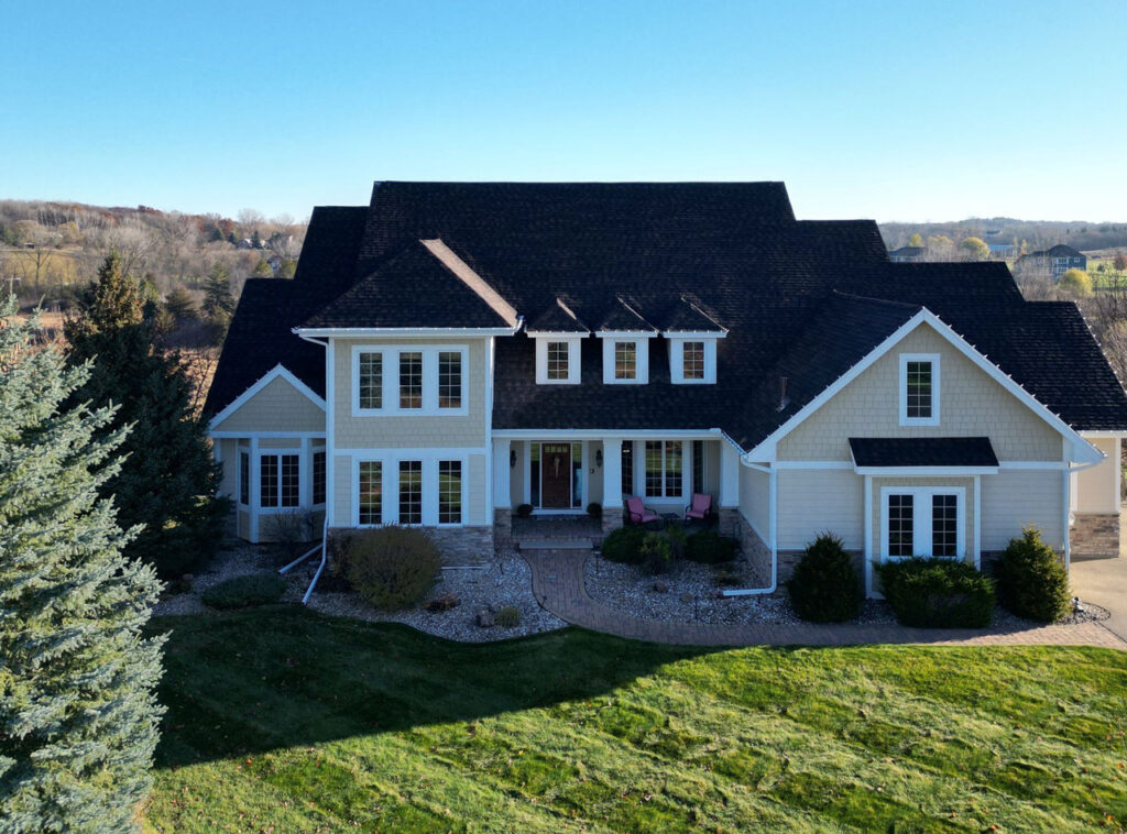 An aerial shot of a large, custom-built home with beige siding and a dark shingle roof, set on a sprawling green lawn.