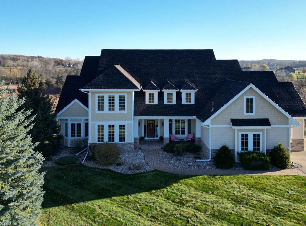 An aerial shot of a large, custom-built home with beige siding and a dark shingle roof, set on a sprawling green lawn.