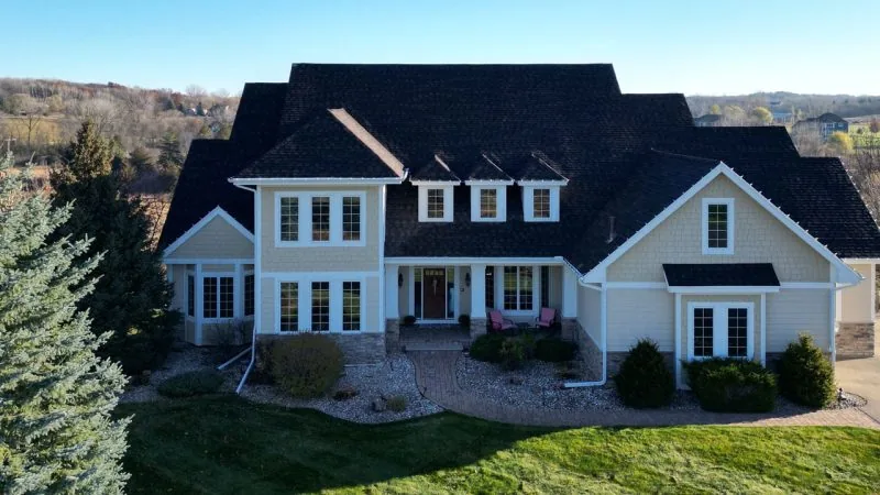 An aerial shot of a large, custom-built home with beige siding and a dark shingle roof, set on a sprawling green lawn.