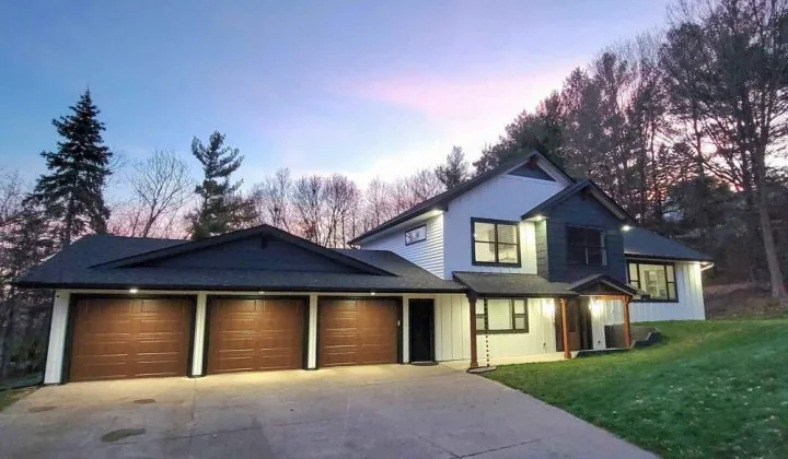 A modern custom home featuring white and dark blue siding is beautifully illuminated by exterior lights against a twilight sky.