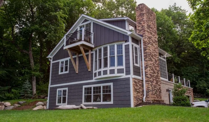 A stylish, multi-level home featuring dark gray horizontal siding, white trim, and a prominent brick chimney, nestled in a lush green landscape.