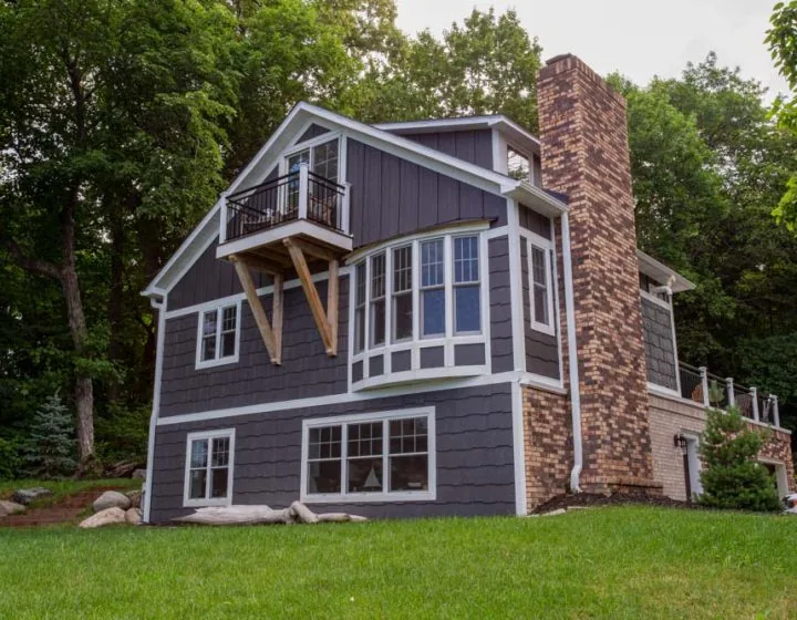 A stylish, multi-level home featuring dark gray horizontal siding, white trim, and a prominent brick chimney, nestled in a lush green landscape.