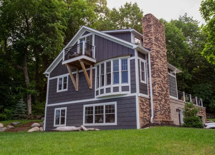 A stylish, multi-level home featuring dark gray horizontal siding, white trim, and a prominent brick chimney, nestled in a lush green landscape.