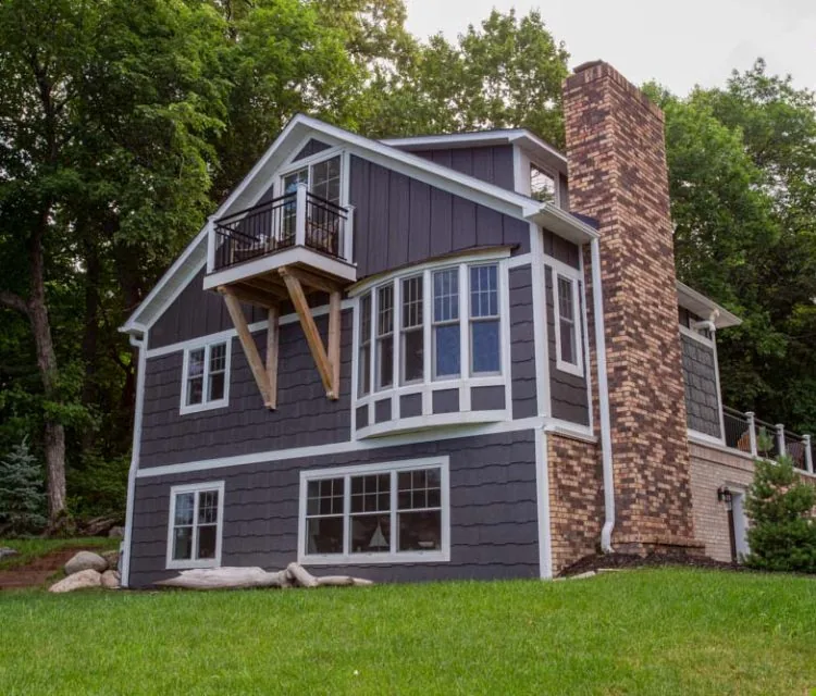 A stylish, multi-level home featuring dark gray horizontal siding, white trim, and a prominent brick chimney, nestled in a lush green landscape.