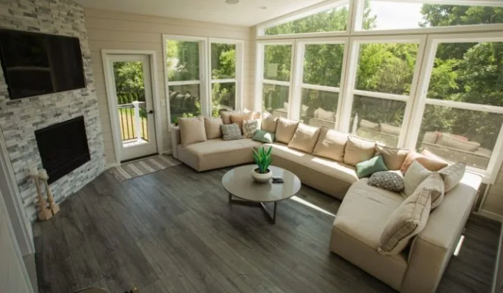 Contemporary sunroom featuring dark wood flooring, a stone fireplace, sectional sofa, and expansive corner windows looking out to nature.