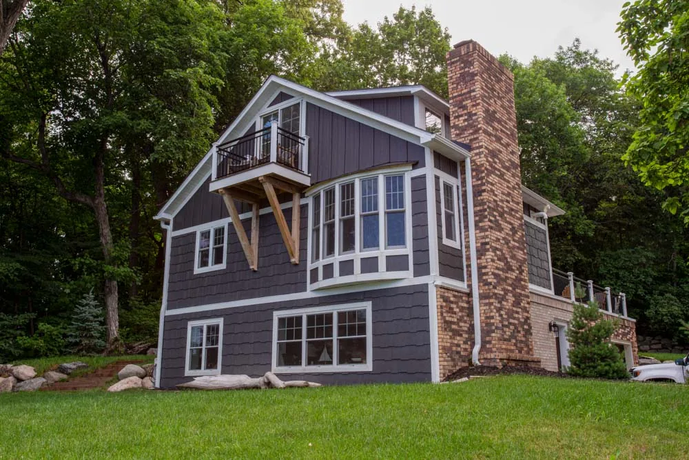 A stylish, multi-level home featuring dark gray horizontal siding, white trim, and a prominent brick chimney, nestled in a lush green landscape.