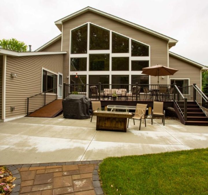Exterior view of a beige home featuring a massive custom geometric window wall overlooking a concrete patio and wooden deck.