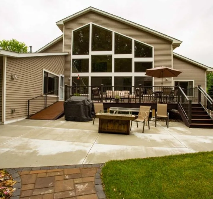 Exterior view of a beige home featuring a massive custom geometric window wall overlooking a concrete patio and wooden deck.