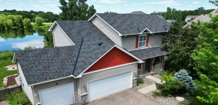 Aerial view of a two-story lakefront home featuring newly installed dimensional asphalt shingles in charcoal gray, complemented by beige siding and red gable accent.