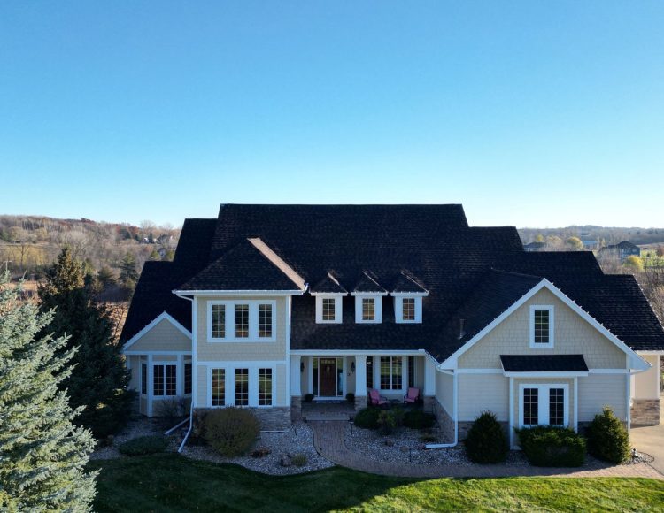 A grand, multi-gabled home with light beige siding, white trim, and a dark shingle roof against a clear blue sky.