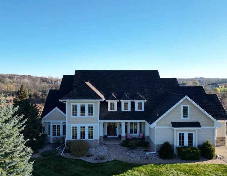 A grand, multi-gabled home with light beige siding, white trim, and a dark shingle roof against a clear blue sky.