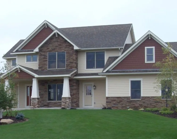 Front elevation of a custom-built home combining cream-colored lap siding, red-brown shingle accents, stone veneer columns, and dark roof shingles with landscaped front yard.