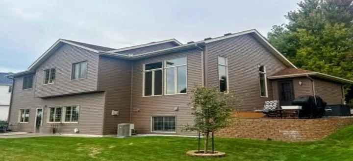 Side-rear view of a modern ranch-style home with rich brown horizontal siding, large picture windows, brick retaining wall, and outdoor patio area surrounded by green grass and trees.