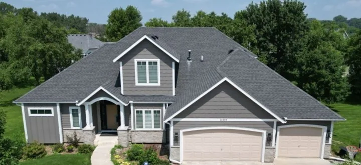 Aerial view of a two-story home with new gray horizontal siding, white trim, dark shingle roof, and dual garage doors surrounded by green landscaping.