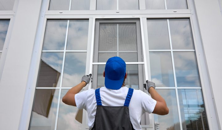 A worker in a blue cap and overalls installs a large, multi-pane white window on the exterior of a modern building.