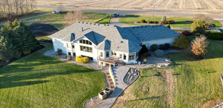 An aerial shot of a sprawling custom home with a gray roof, set on a large grassy lot next to open fields.
