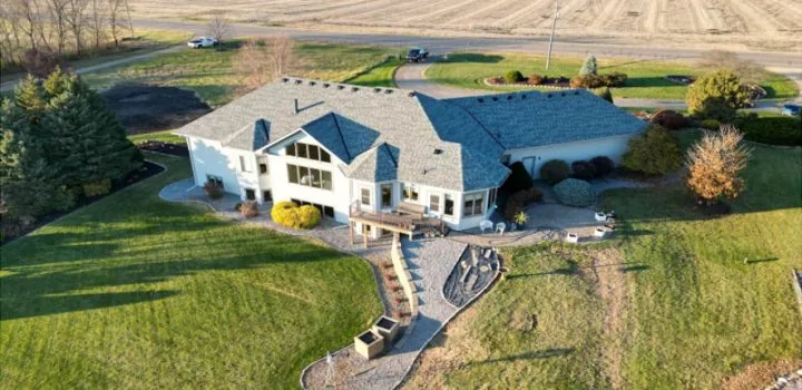 An aerial shot of a sprawling custom home with a gray roof, set on a large grassy lot next to open fields.