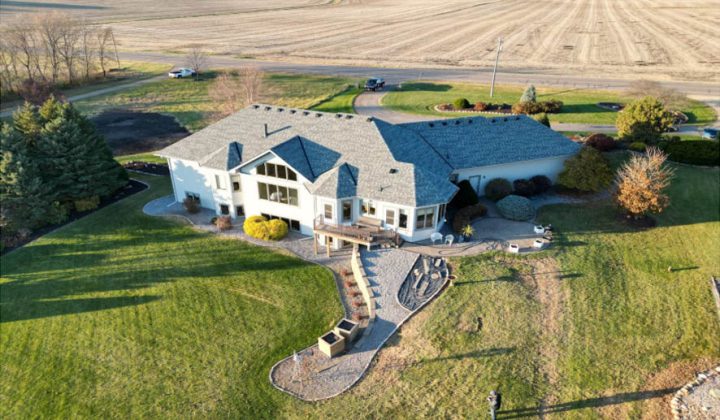 An aerial shot of a sprawling custom home with a gray roof, set on a large grassy lot next to open fields.