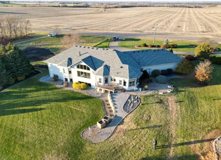 An aerial shot of a sprawling custom home with a gray roof, set on a large grassy lot next to open fields.
