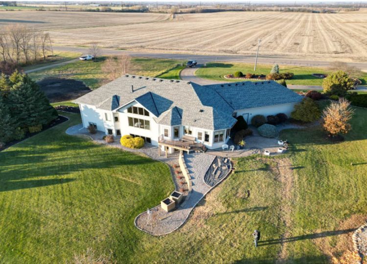 An aerial shot of a sprawling custom home with a gray roof, set on a large grassy lot next to open fields.