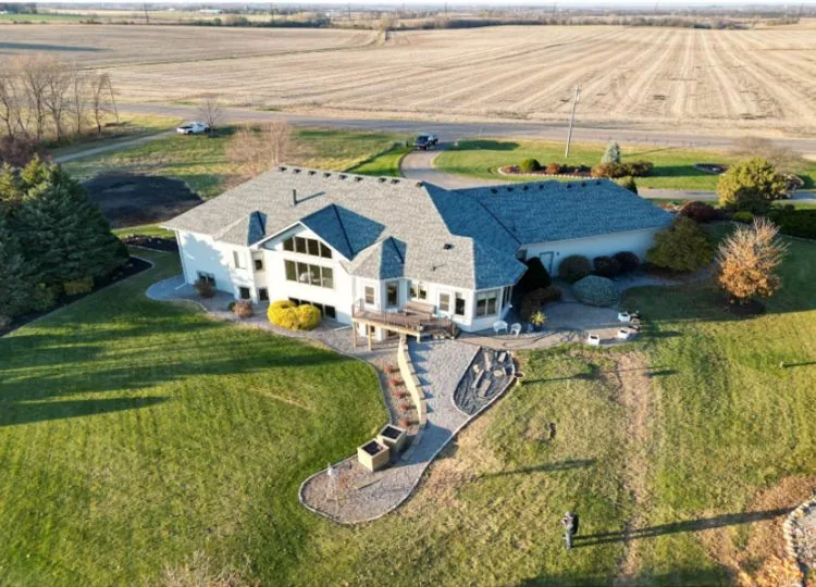 An aerial shot of a sprawling custom home with a gray roof, set on a large grassy lot next to open fields.