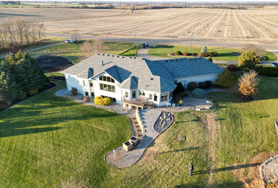 An aerial shot of a sprawling custom home with a gray roof, set on a large grassy lot next to open fields.