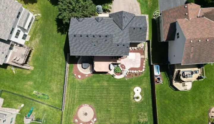 An overhead shot of a suburban home with a gray roof, surrounded by a large green lawn and custom hardscaping.