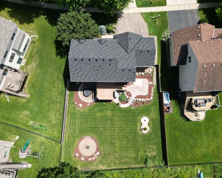An overhead shot of a suburban home with a gray roof, surrounded by a large green lawn and custom hardscaping.