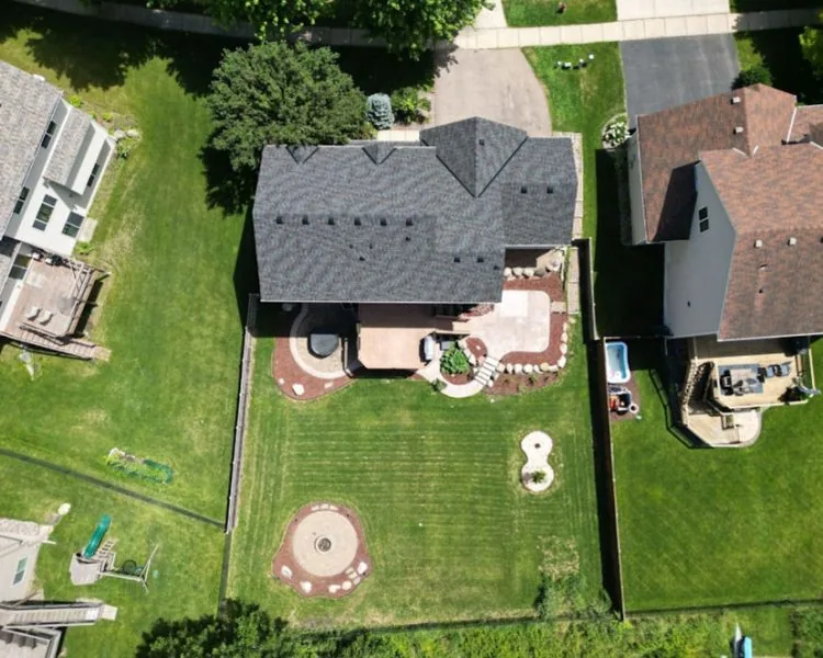 An overhead shot of a suburban home with a gray roof, surrounded by a large green lawn and custom hardscaping.