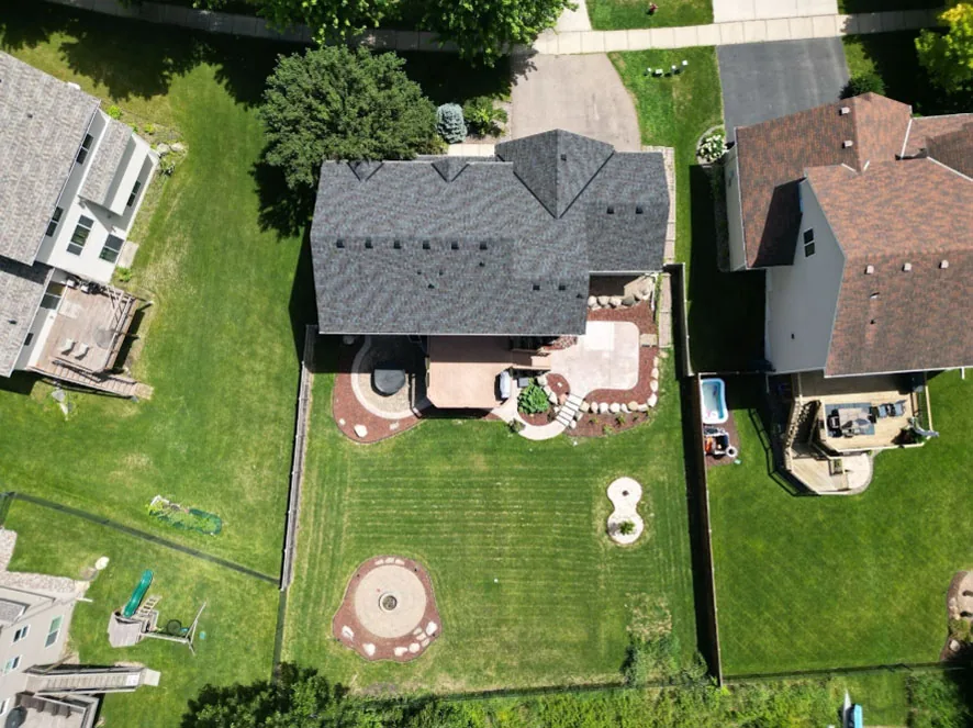 An overhead shot of a suburban home with a gray roof, surrounded by a large green lawn and custom hardscaping.
