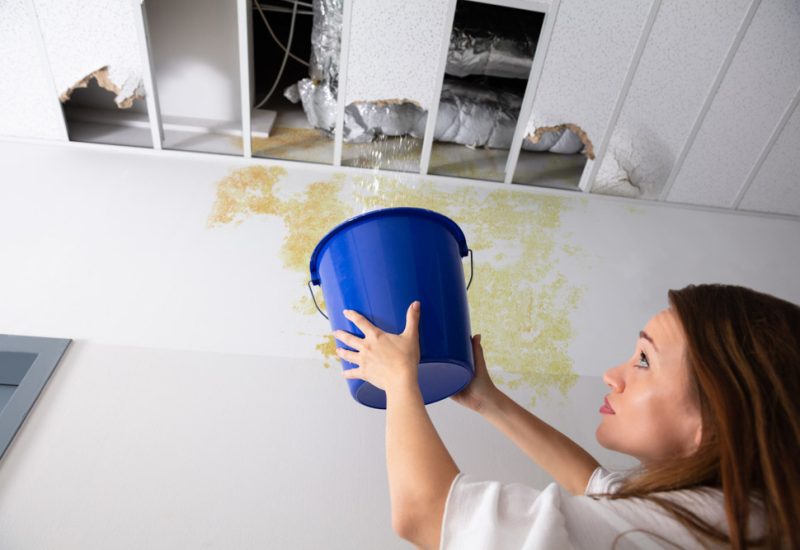 Worried Woman Holding A Blue Bucket Under The Leak Ceiling In Corridor