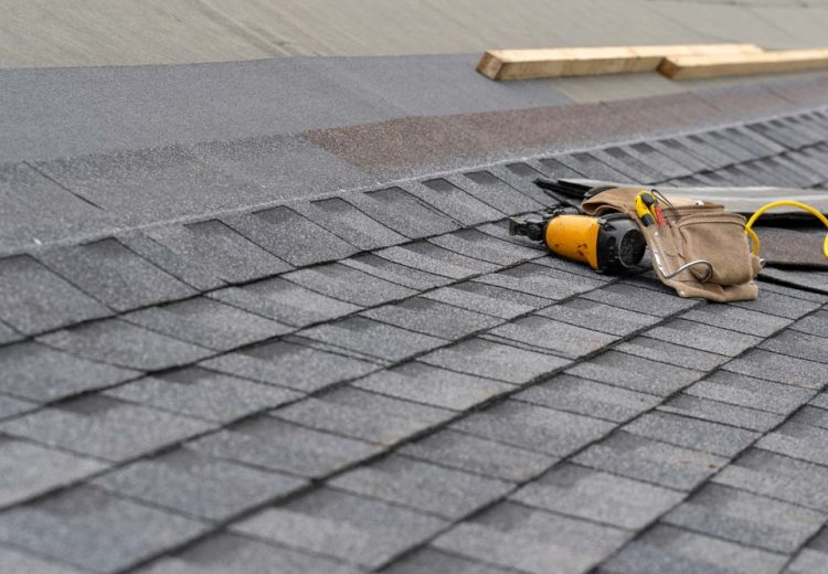 Three roofers work together to install asphalt shingles on a house, using nail guns and safety ropes.