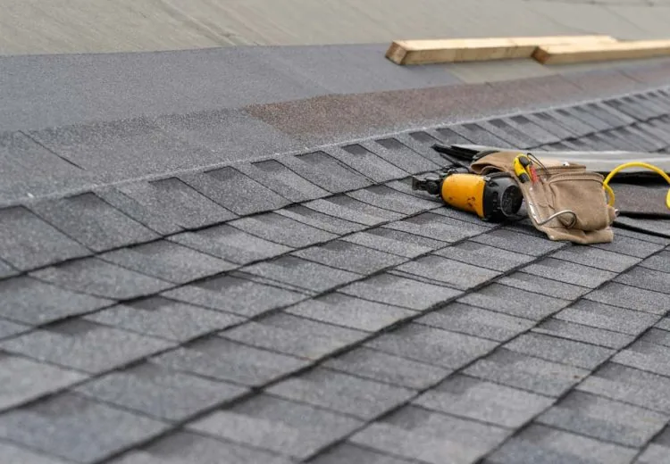 Photo of toolbelt with instrument and nail gun lying on asphalt or bitumen shingle on top of the new roof under construction residential house or building