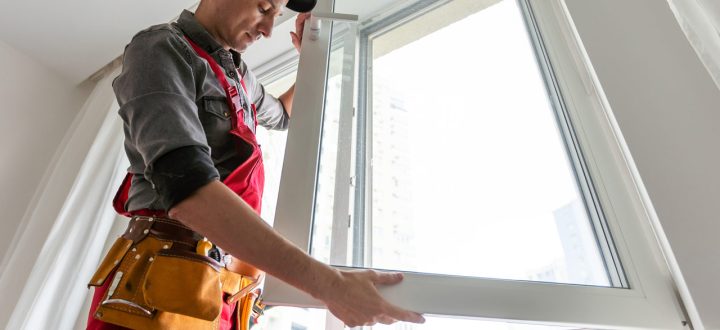 A skilled window installer in red overalls carefully fits a new white window frame into place inside a bright room.