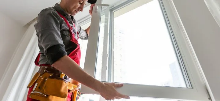 A skilled window installer in red overalls carefully fits a new white window frame into place inside a bright room.
