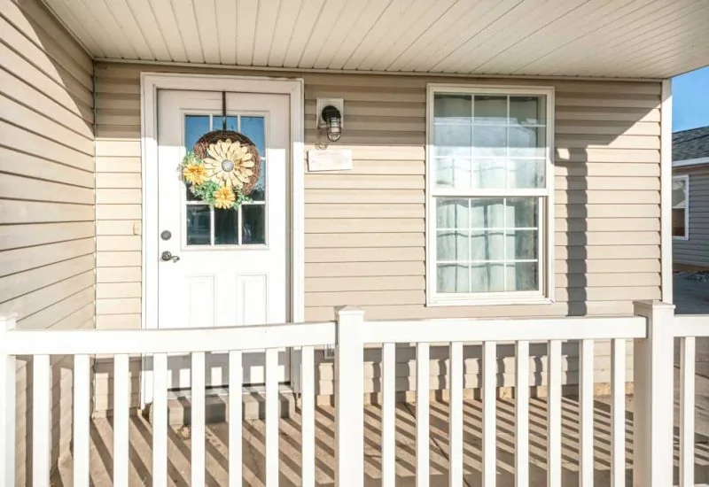 Porch of a house with beige vinyl wood siding and railing. There is a white front door with wreath on top of the glass panels near the single hung window with curtain.