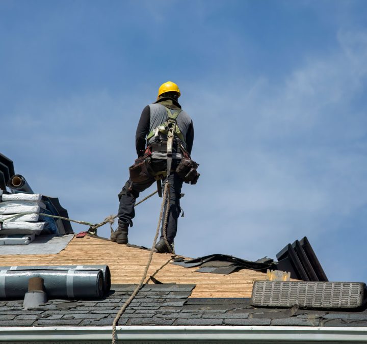 A roofer in safety gear works on a roof under a clear blue sky, installing new shingles.