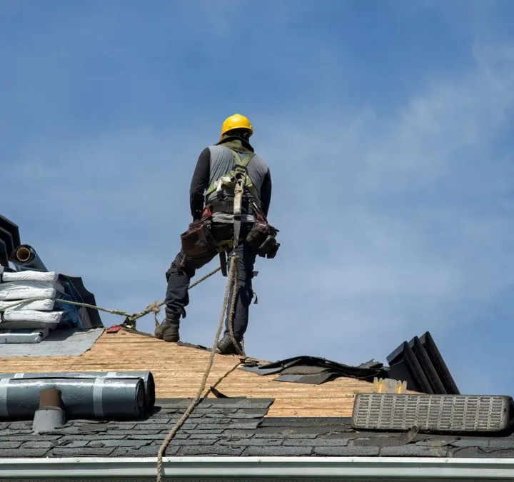 A roofer in safety gear works on a roof under a clear blue sky, installing new shingles.