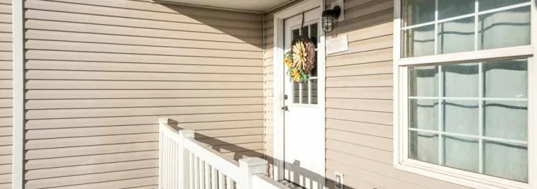 Front door exterior of a house with vinyl wall sidings. There is a white wooden railings at the front of a door with glass panel and wreath beside the double hung window.