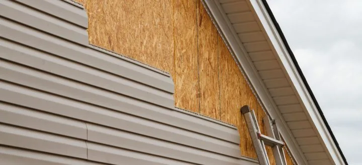 A close-up view of beige horizontal siding being installed on a home, showing exposed OSB sheathing and a ladder.