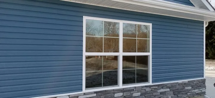 A close-up of a home's exterior featuring blue horizontal siding, a white-framed window, and a stone veneer foundation.
