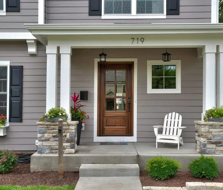 The inviting front entrance of a home with gray siding, a wooden door, white trim, and decorative stone pillars.